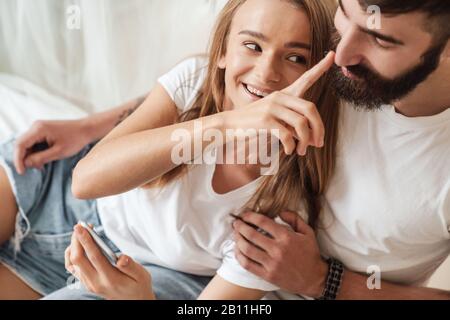 Immagine di felice giovane coppia che fa divertimento e usando il cellulare mentre abbracciando in stanza luminosa a casa Foto Stock