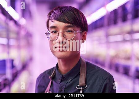 Ritratto di giovane asiatico in occhiali guardando la macchina fotografica mentre si sta in piedi nella pianta Foto Stock