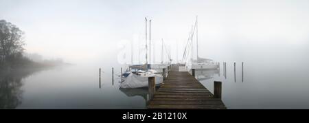 Mattina d'autunno sul lago Chiemsee, molo e barche a vela in nebbia, Baviera, Germania Foto Stock