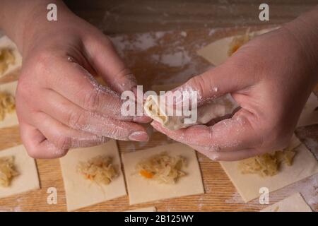 Una donna scolpisce gnocchi e ravioli da quadrati di pasta e cavolo. Pannello di taglio compensato, setaccio di farina di legno e perno di laminazione di legno - attrezzi per Foto Stock