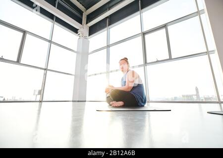 Giovane femmina disable con le gambe incrociate che fanno esercizio di yoga sul tappetino in palestra Foto Stock