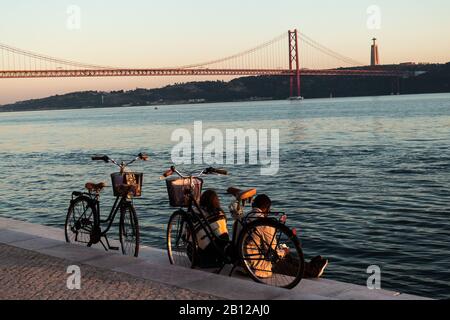 Il ponte del 25 aprile è un ponte sospeso attraverso il fiume Tajo in Portogallo, che collega Lisbona e Almada. 2 ciclisti appoggiati sulla riva. Foto Stock