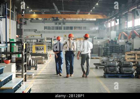 Vista posteriore di tre lavoratori di impianti industriali che camminano lungo una grande fabbrica Foto Stock