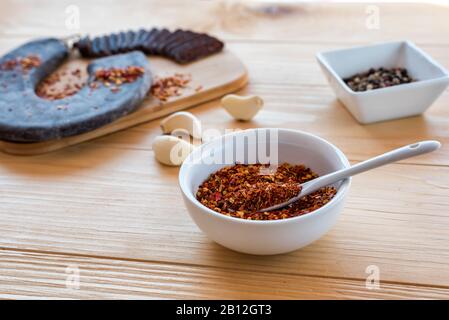 Peperoncino piccante e caldo Tritato secco in ciotola bianca - Tucana ljuta paprika di fronte a speciale salsiccia stirata, garlick e una ciotola di pepe misto Foto Stock