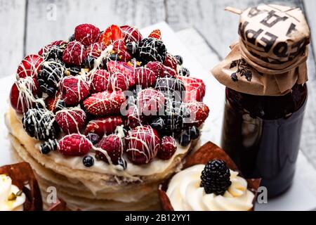 Torta di frittelle con un berretto di frutti di bosco inzuppato in cioccolato bianco su un tavolo bianco di legno con alcuni muffin belgi e una bottiglia di sciroppo Foto Stock