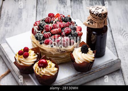 Torta di frittelle con un berretto di frutti di bosco inzuppato in cioccolato bianco su un tavolo bianco di legno con alcuni muffin belgi e una bottiglia di sciroppo Foto Stock