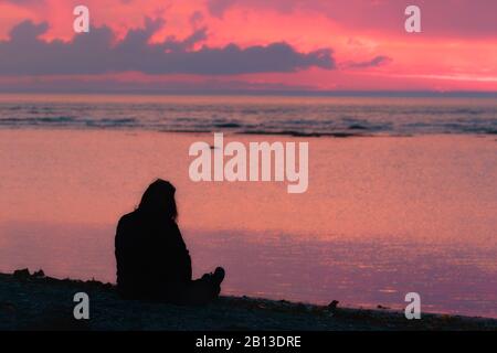 Silhouette di donna seduta sulla spiaggia e guardando il tramonto rosa sopra l'acqua Foto Stock