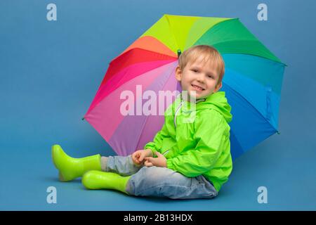 Un bambino caucasico si siede in una giacca antivento e stivali di gomma sotto un ombrello - un arcobaleno, su uno sfondo blu. Concetto su tempo e stagioni Foto Stock