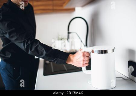 La vista del taglio dell'uomo nei vestiti neri tiene il bollitore elettrico in mano. L'acqua si riversa dal rubinetto. bollitore bianco intelligente per il riscaldamento dell'acqua. Giovane uomo in cucina wan Foto Stock