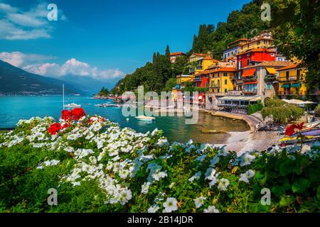 Giardino fiorito sulla riva del lago e splendida vista con edifici colorati. Barche e motoscafi ormeggiati nella baia, lago di Como, Varenna, Italia, Europa Foto Stock