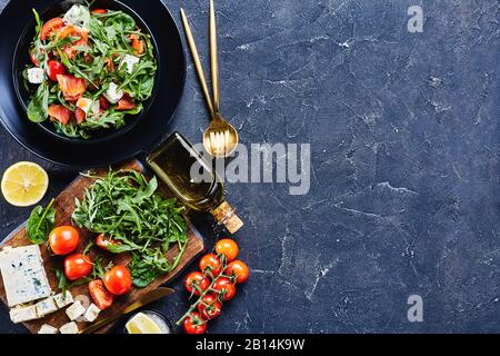 primo piano di deliziosa insalata di pomodoro di rucola di salmone blu in una ciotola nera con forchetta dorata, coltello, cucchiaio e ingredienti, vista orizzontale Foto Stock