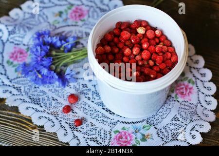 Secchiello bianco pieno di fragole deliziose. Tre bacche di fragola selvatica si trovano su un bouquet di tovaglioli di tutolo in maglia di fiori di mais. Foto Stock