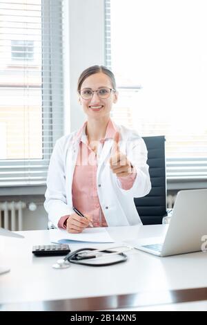 Medico donna dando Thumbs up seduto sulla scrivania nel suo ufficio Foto Stock