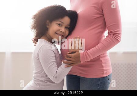 Cute bambina nera che abbraccia e tocca la pancia della madre incinta Foto Stock