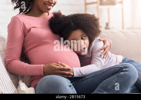 La bambina eccitata tocca e ascolta la pancia della madre incinta Foto Stock