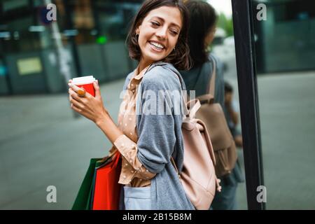 Felice giovane donna bere togliere il caffè e camminare con le borse dopo lo shopping in città. Foto Stock