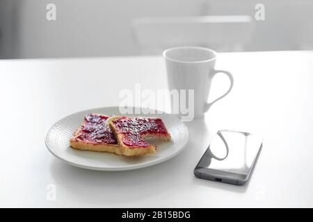 La colazione è a tavola. Una tazza di caffè e toast con marmellata di ciliegie accanto al telefono. Foto Stock