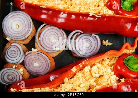 Primo piano di due peperoni rossi cotti al forno riempiti di peperoncino piccante feta e pomodori con anelli di cipolla sul vassoio del forno Foto Stock