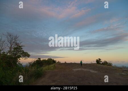 Lady fotografo in distanza che appare piccolo sotto il cielo crepuscolo, a piedi dal bordo delle scogliere con un tramonto e la luna dietro di lei Foto Stock