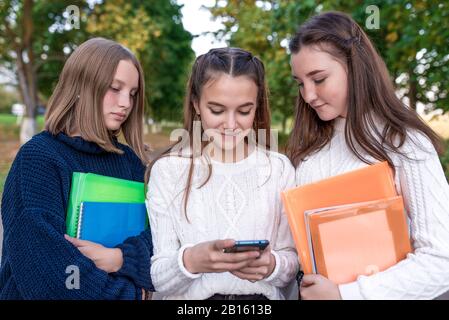 Tre ragazze, scolaresche, adolescenti 12-15 anni, parco estivo, guardare video telefono, cartella notebook abstracts con libri di testo. Scuola pausa riposo Foto Stock