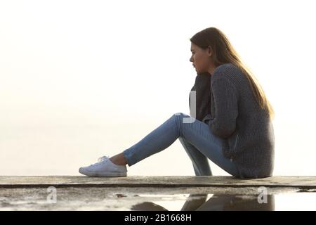 Vista laterale ritratto di una ragazza triste che guarda lontano da sola seduta all'aperto contemplando orizzonte Foto Stock