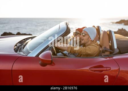 Coppia gioiosa godendosi le vacanze, guidando insieme l'automobile convertibile sulla costa rocciosa dell'oceano su un tramonto. Felice vacanza, amore e concetto di viaggio Foto Stock