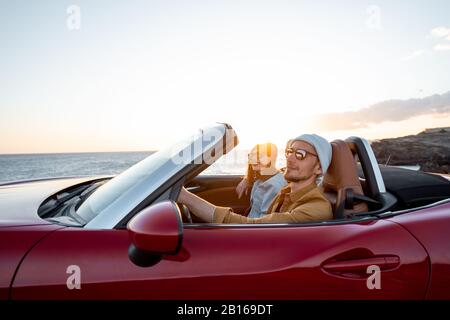 Coppia gioiosa godendosi le vacanze, guidando insieme l'automobile convertibile vicino all'oceano su un tramonto. Felice vacanza, amore e concetto di viaggio Foto Stock