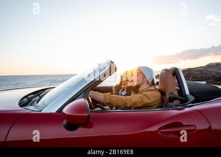 Coppia gioiosa godendosi le vacanze, guidando insieme l'automobile convertibile vicino all'oceano su un tramonto. Felice vacanza, amore e concetto di viaggio Foto Stock