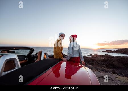 Ritratto di una bella coppia vestito casualmente in cappelli seduti insieme su una macchina durante un tramonto, viaggiando vicino all'oceano. Buone vacanze e viaggi in auto