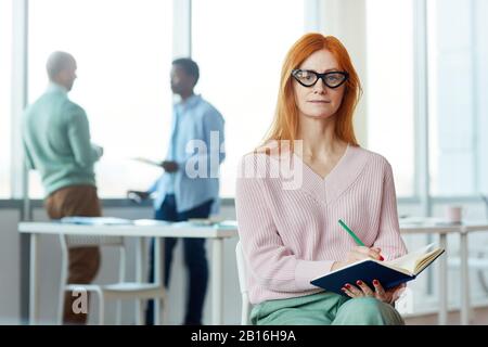 Ritratto di businesswoman rosso guardando la fotocamera e tenendo pianificatore mentre posa in ufficio impostazione, copia spazio Foto Stock