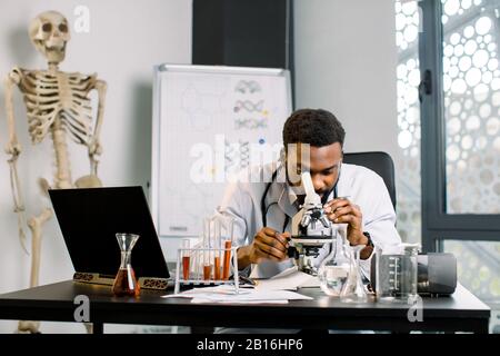 Un bel tecnico africano, medico, microbiologo, in laboratorio guardando al microscopio, lavorando alla ricerca medica, seduto al tavolo wi Foto Stock