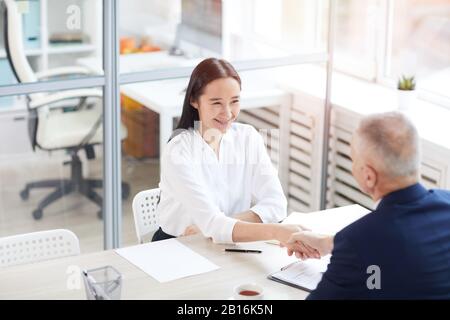 Ritratto ad alto angolo di giovane donna d'affari asiatica sorridente felicemente mentre scuotendo le mani con l'uomo anziano attraverso il tavolo in ufficio, copia spazio Foto Stock