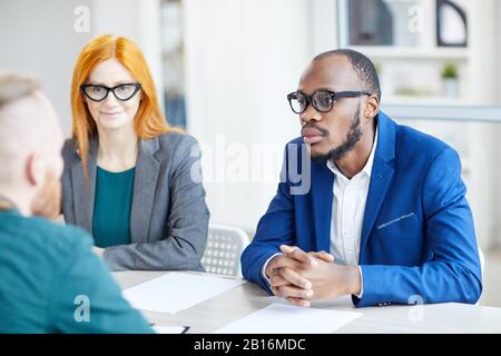 Ritratto dell'uomo d'affari afroamericano contemporaneo che ascolta il candidato durante l'intervista di lavoro in ufficio Foto Stock