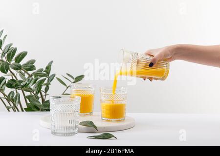 vista dal taglio corto della donna che versa un delizioso frullato giallo in vetro sulla superficie bianca vicino alla pianta verde Foto Stock