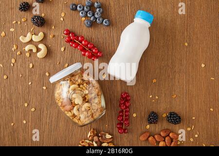 Vista dall'alto di frutti di bosco, noci, vasetto di granola e bottiglia di yogurt su sfondo di legno Foto Stock