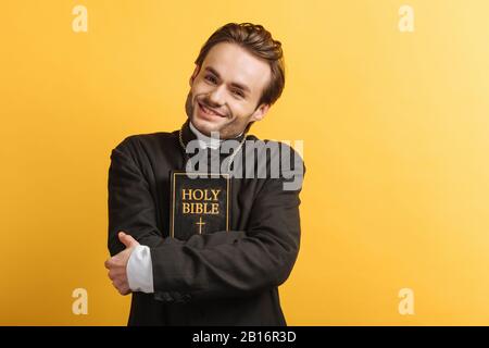 felice sacerdote cattolico guardando la macchina fotografica mentre abbracciando la bibbia isolato su giallo Foto Stock