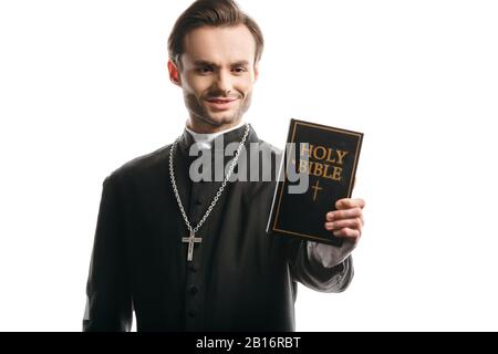 giovane sacerdote cattolico sorridente mentre tiene la sacra bibbia isolata su bianco Foto Stock