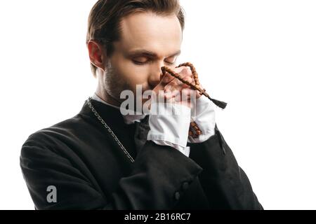 giovane sacerdote cattolico concentrato che prega con occhi chiusi mentre tiene perline di legno rosario vicino al viso isolato su bianco Foto Stock