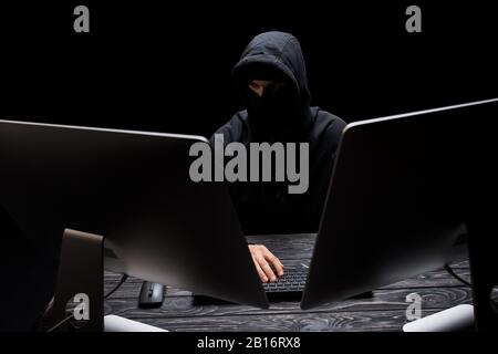 hacker in maschera seduto vicino a monitor di computer isolato su nero Foto Stock