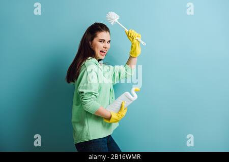 casalinga irritata tenendo lo stantuffo in mano sollevata e gridando su sfondo blu Foto Stock