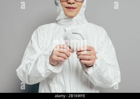 vista ritagliata dell'epidemiologo in tuta hazmat con maschera respiratore su sfondo grigio Foto Stock
