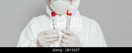 vista ritagliata dell'epidemiologo in tuta e maschera respiratore per il contenimento di provette con campioni di sangue isolati su shot panoramici grigi Foto Stock