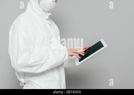 vista ritagliata dell'epidemiologo in tuta hazmat e maschera respiratoria con tablet digitale con schermo vuoto su sfondo grigio Foto Stock