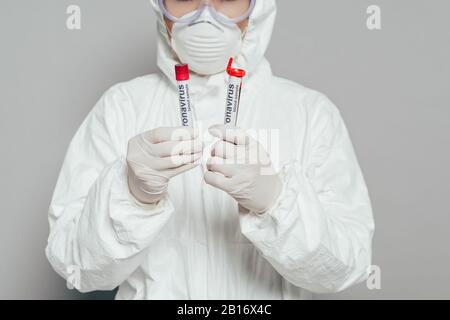 vista ritagliata dell'epidemiologo in tuta e maschera respiratore per il contenimento di provette con campioni di sangue su sfondo grigio Foto Stock
