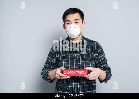 giovane uomo asiatico in maschera respiratore che mostra la carta di avvertimento con iscrizione coronavirus su sfondo grigio Foto Stock