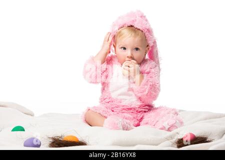 Bambino che indossa il costume di coniglio, mettendo l'uovo di pasqua in bocca, toccando la testa su coperta isolata su bianco Foto Stock
