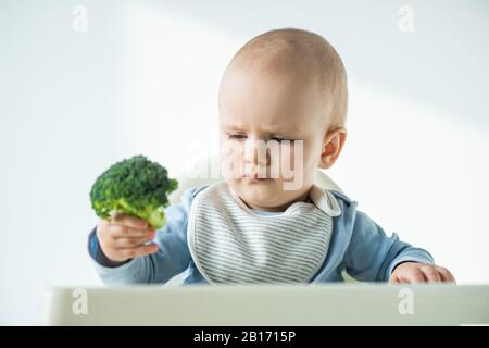 Fuoco selettivo del pezzo di broccoli che tiene il bambino mentre si siede sulla sedia di alimentazione su sfondo bianco Foto Stock