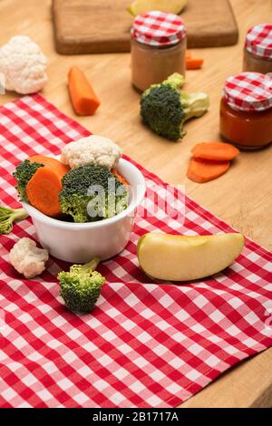 Verdure organiche e mela su tovagliolo vicino vasi di alimenti per bambini su sfondo di legno Foto Stock