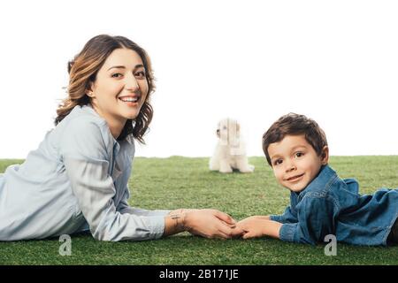 Fuoco selettivo di madre sorridente e del figlio che giace sull'erba e sul cucciolo di Havanese sullo sfondo isolato su bianco Foto Stock