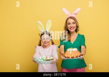 allegra madre con orecchie bunny che tiene la torta di pasqua vicino a figlia con uova di pollo dipinte isolato su giallo Foto Stock